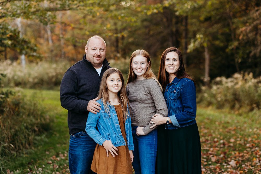 A family stands together outdoors on an autumn day with fallen leaves and foliage in the background.