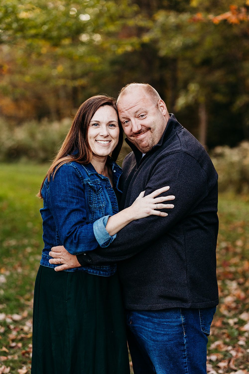 A couple embraces outdoors in an autumn setting with fallen leaves and trees in the background.