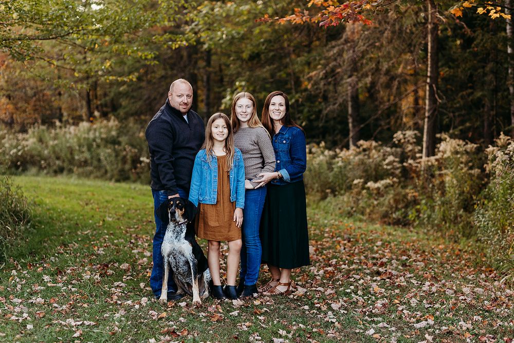 A family and their grey dog stand together outdoors on fallen autumn leaves with colorful trees in the background.