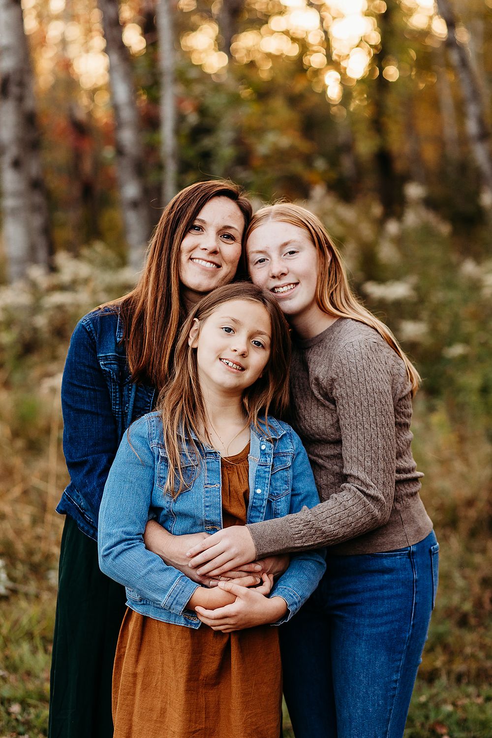 A family portrait outdoors with autumn trees and bokeh lighting in the background.