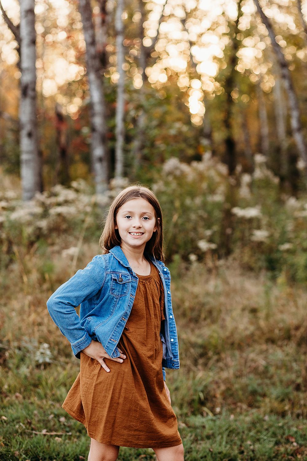 A child wearing a denim jacket and brown dress poses outdoors in a birch forest during golden hour sunset.