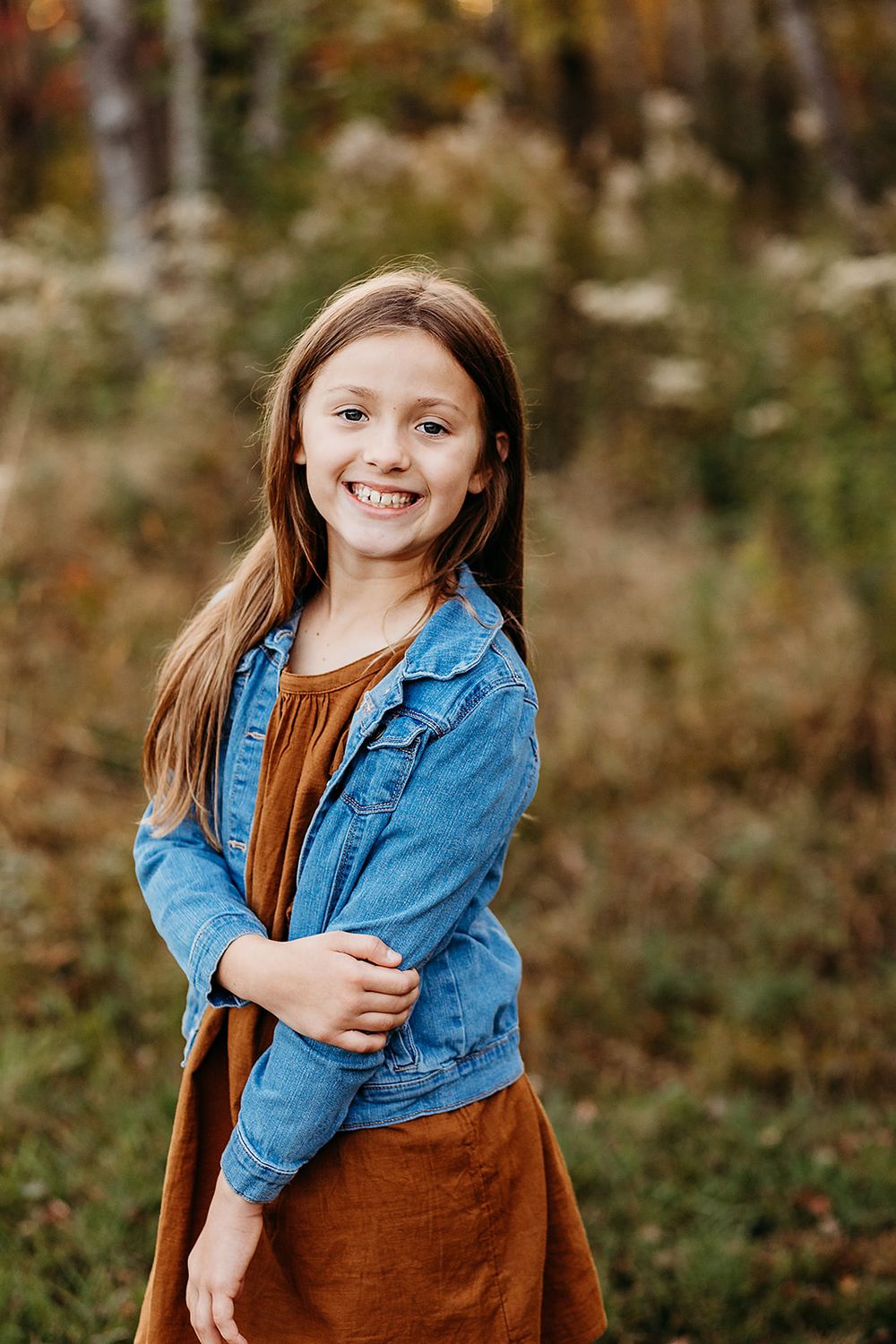 Cheerful youth wearing denim jacket and brown dress poses outdoors in autumn setting.