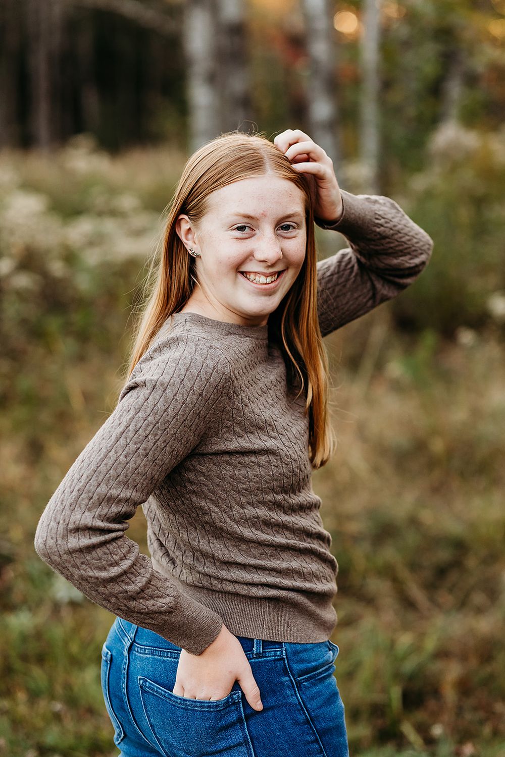 Casual portrait of young person in brown sweater and jeans smiling in natural outdoor setting.