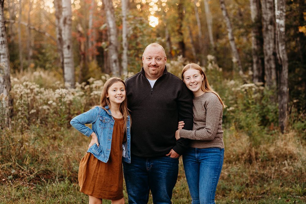 Three people in casual clothing stand together smiling in an autumn forest with birch trees at sunset.