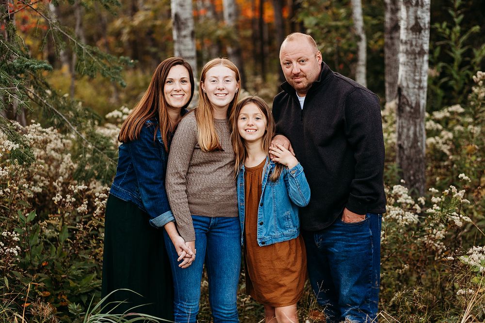 A group stands together outdoors in casual attire against an autumn forest backdrop with birch trees and fall foliage.