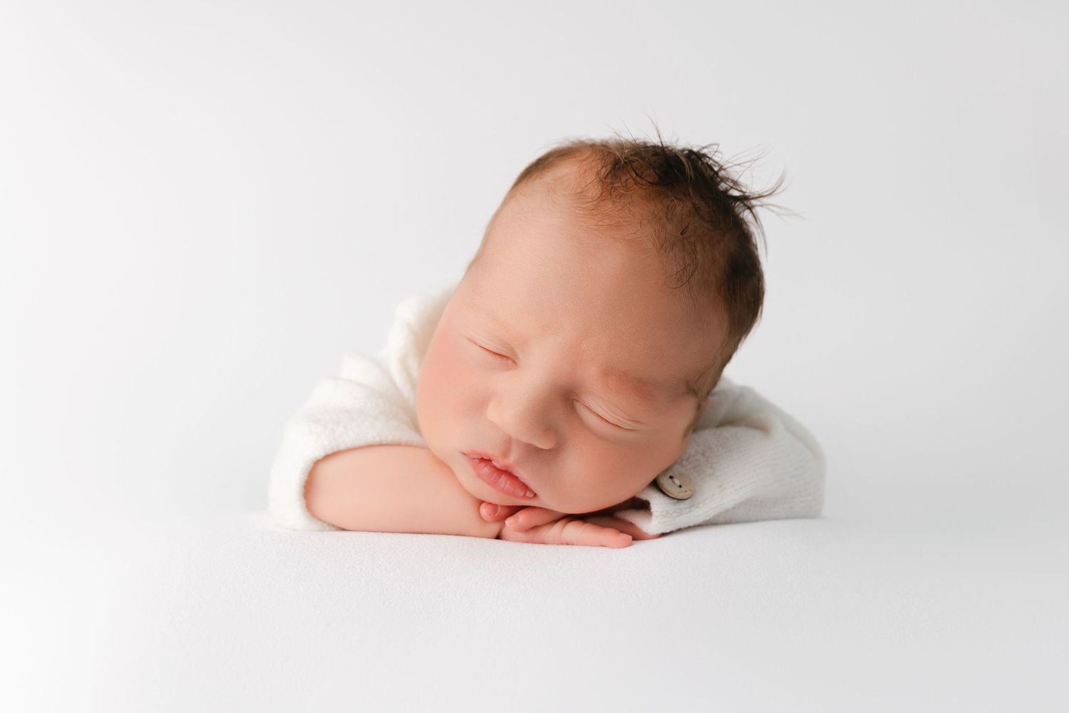 A newborn baby sleeps peacefully on a white backdrop with hands tucked under their cheek.