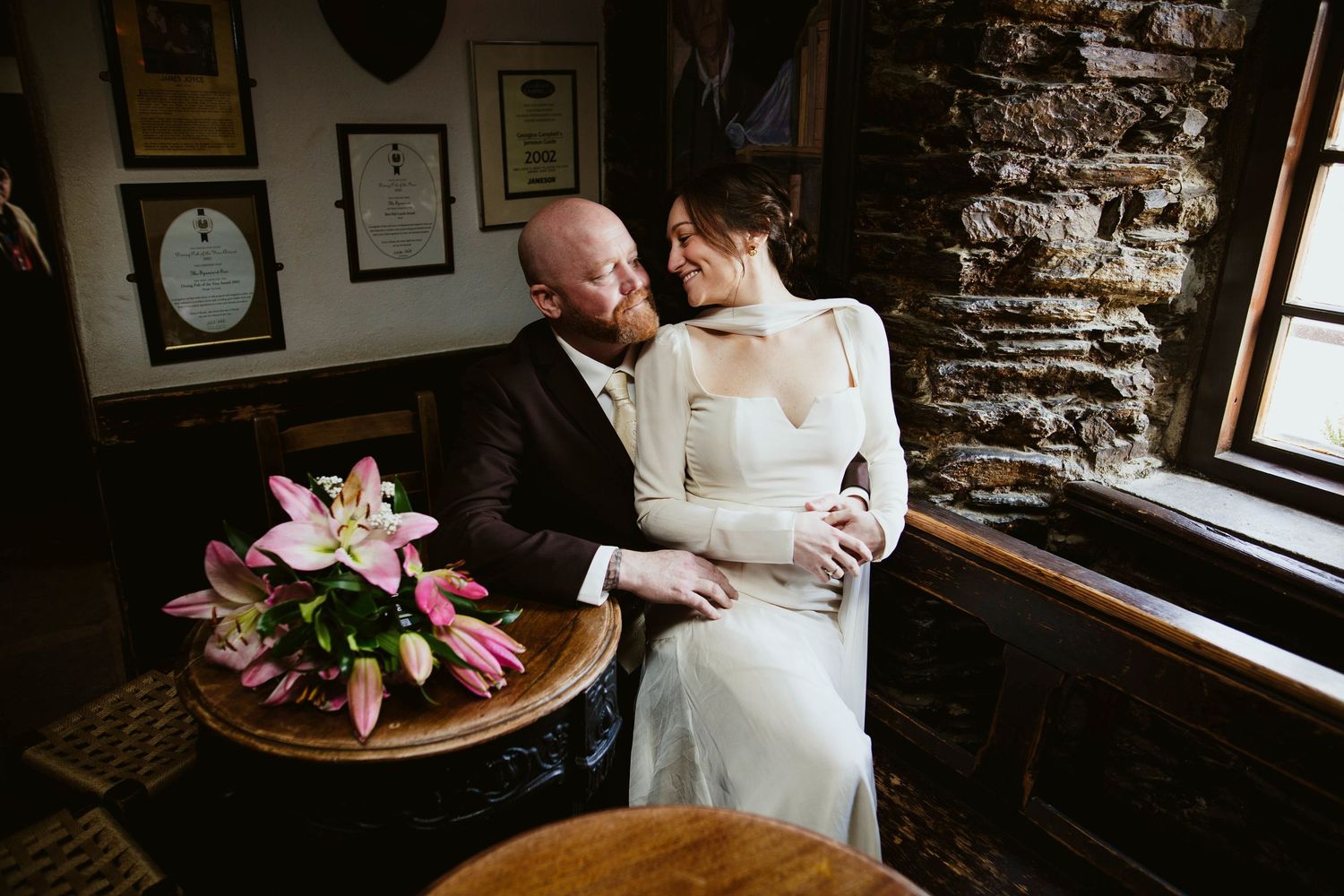 A romantic wedding moment in Kinsale captured in a rustic pub interior with stone walls and a pink lily bouquet on a wooden table.