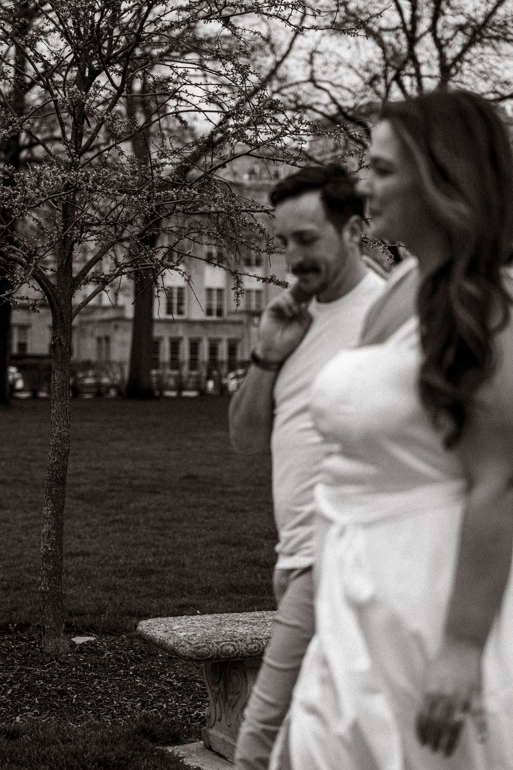 Black and white photo of a couple in a park with leafless trees and buildings in the background.