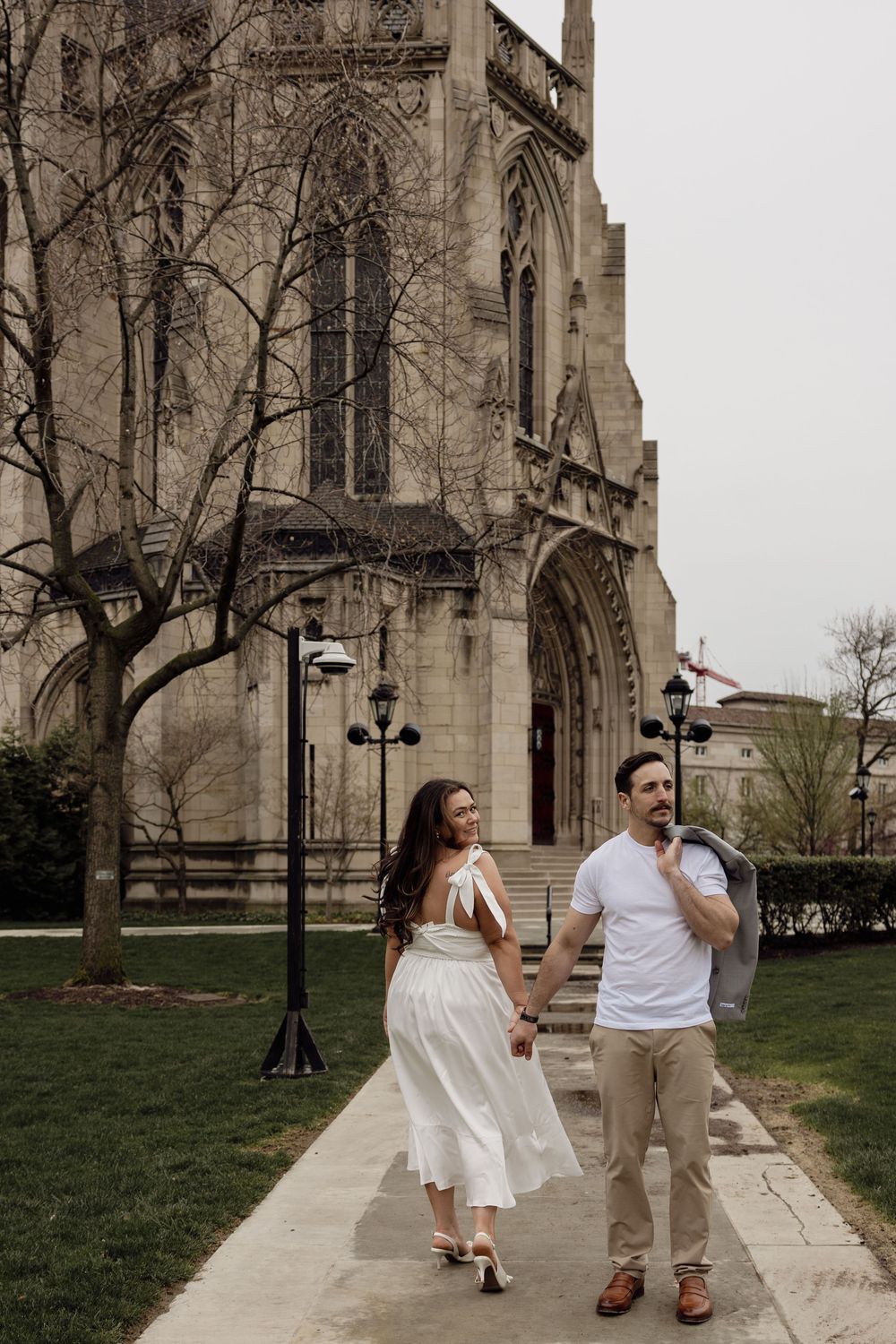 A couple walks hand in hand along a pathway beside a Gothic-style cathedral with ornate architecture and lamp posts.