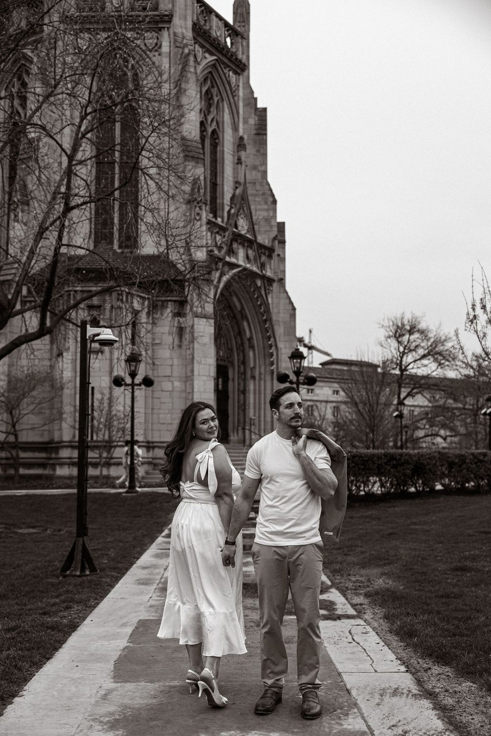 A couple strolls together along a stone path outside a Gothic cathedral in black and white photography.