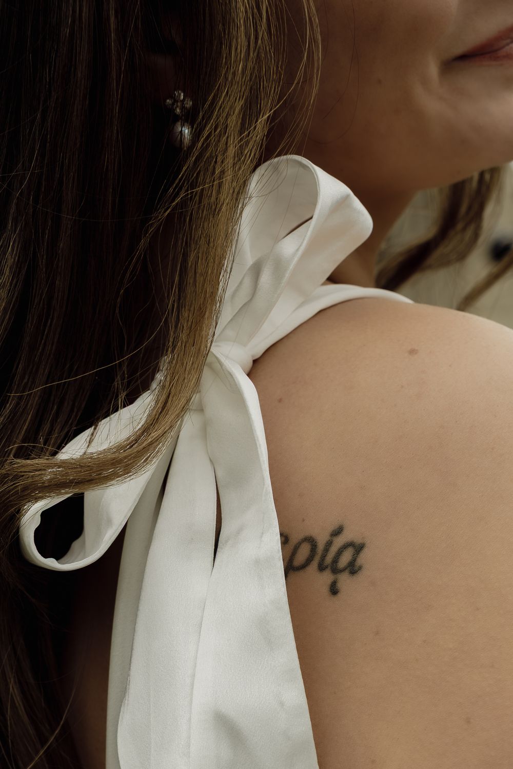 A delicate white bow rests on the shoulder next to a small cursive tattoo against fair skin.