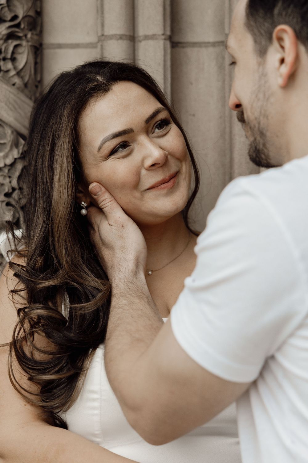 A romantic couple shares an intimate moment against an ornate stone wall background as the man gently touches her cheek.