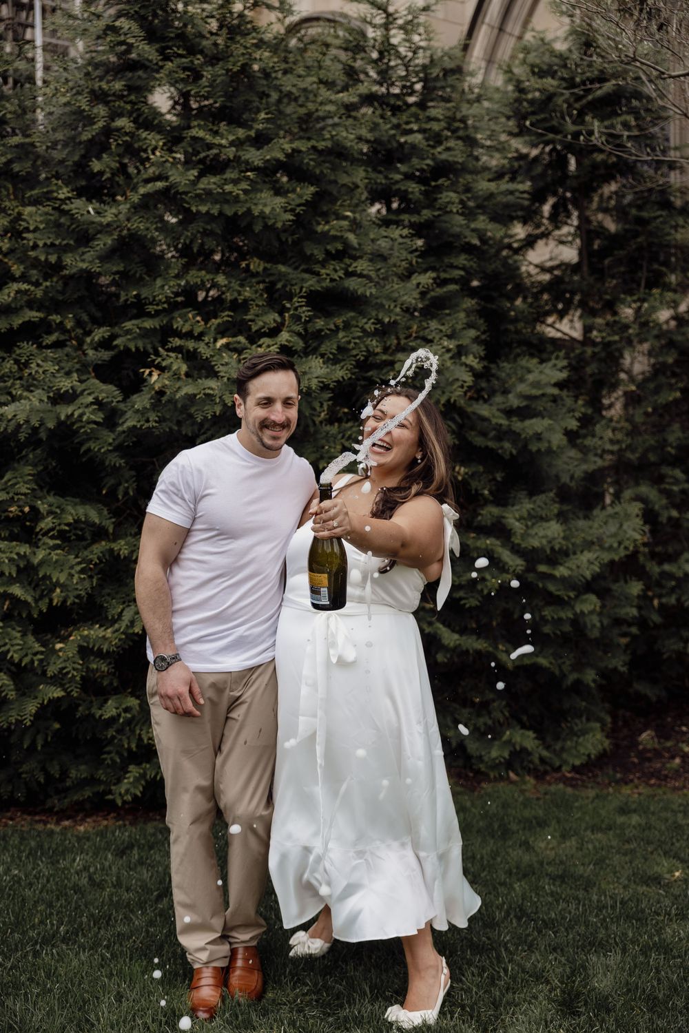 A couple celebrates outdoors by spraying champagne in front of evergreen trees.