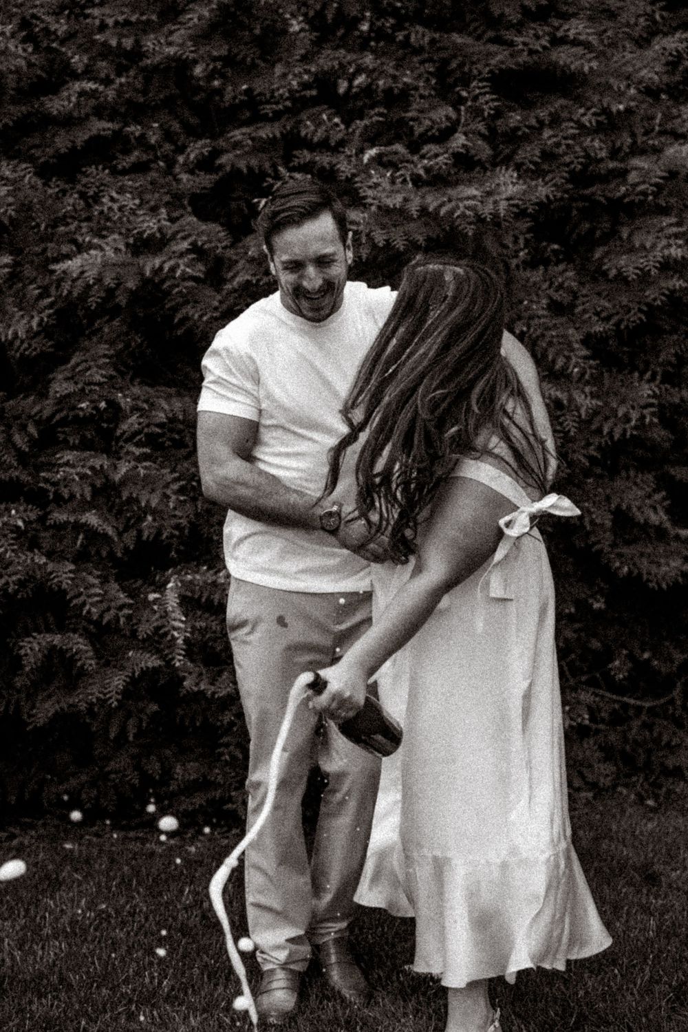 A couple in casual white outfits shares a candid romantic moment in a garden with lush greenery in the background.