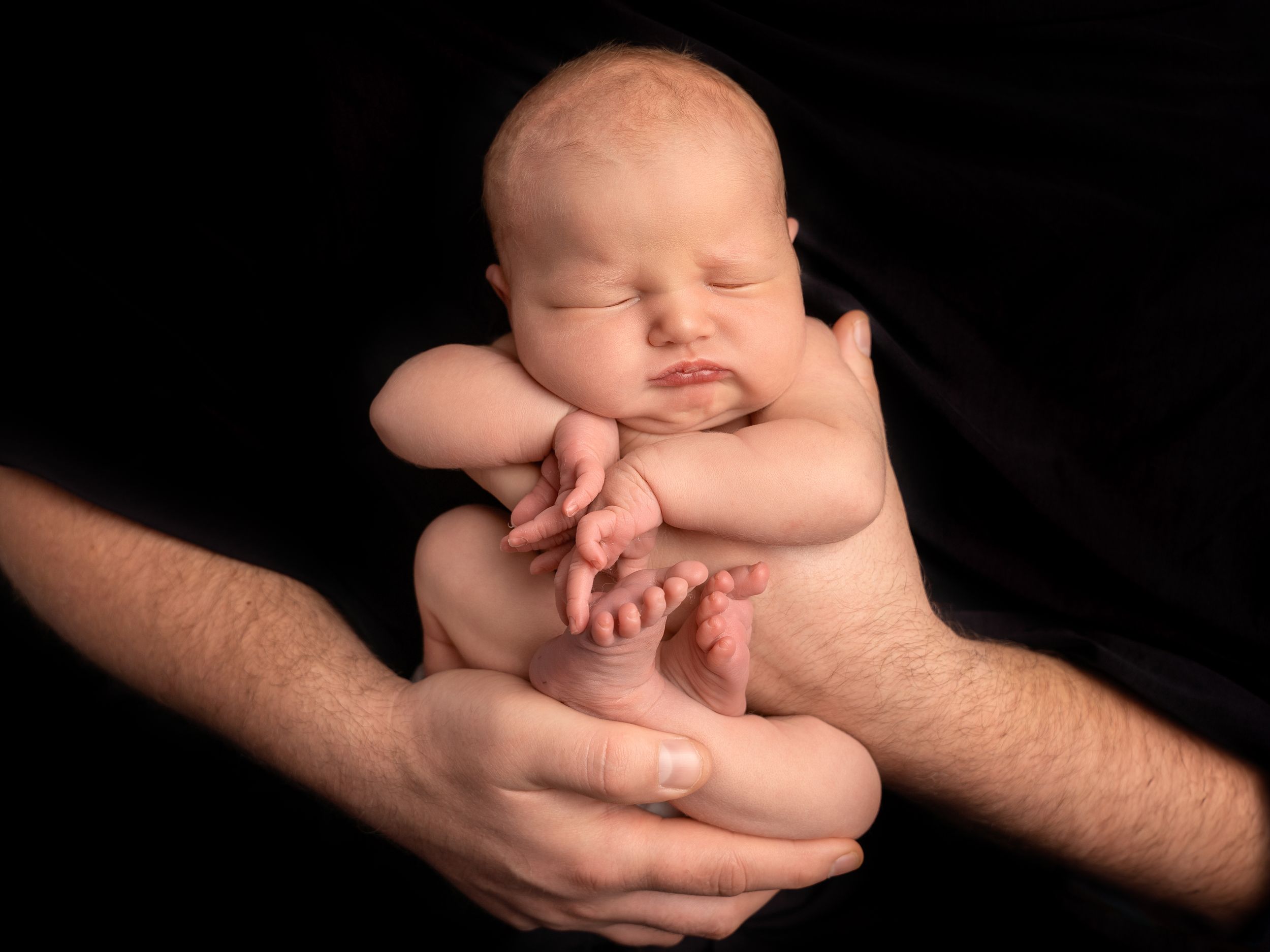 Baby girl held in dad's hands on black backdrop, natural photography with hands, feet and face details at newborn photoshoot in Doncaster.