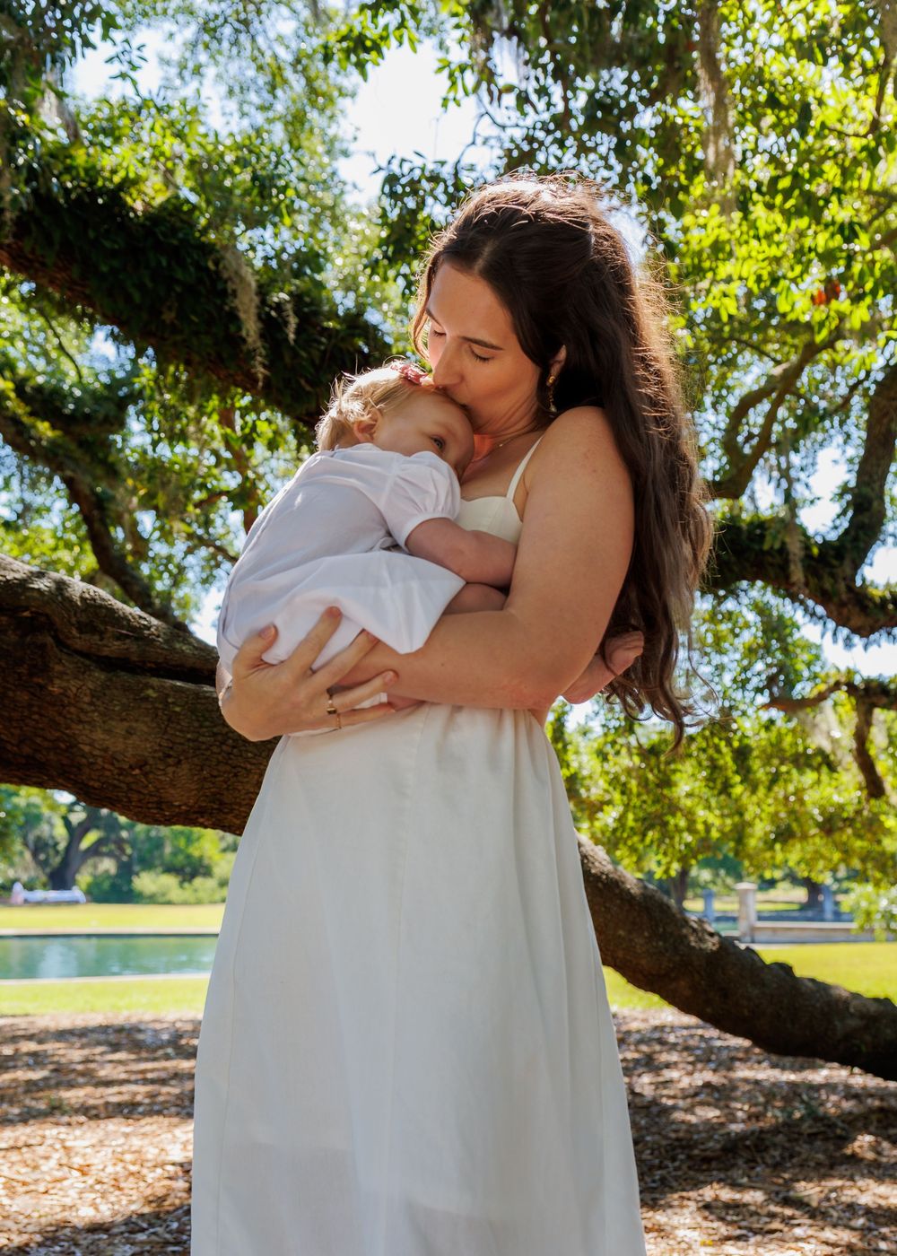 A mother in a white dress tenderly holds and kisses her baby outdoors under a large oak tree in a sunny park.