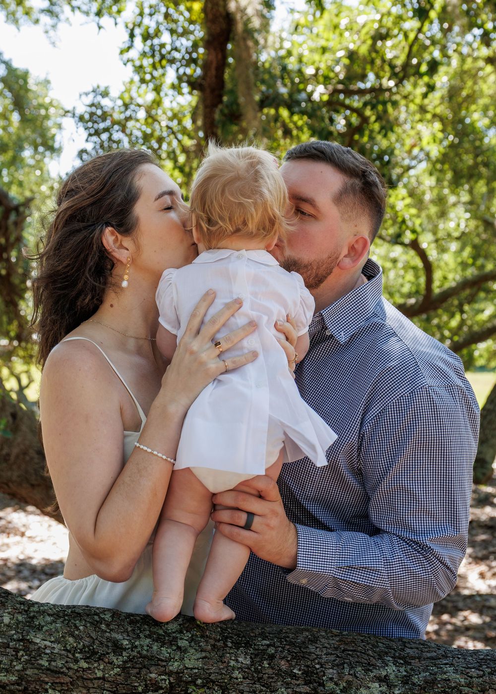A couple kisses their blonde toddler on the cheeks outdoors among lush green trees in a warm family portrait.