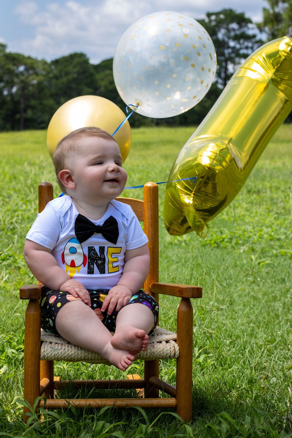 A baby in a bowtie sits in a wooden chair with gold and white balloons for a first birthday photoshoot.