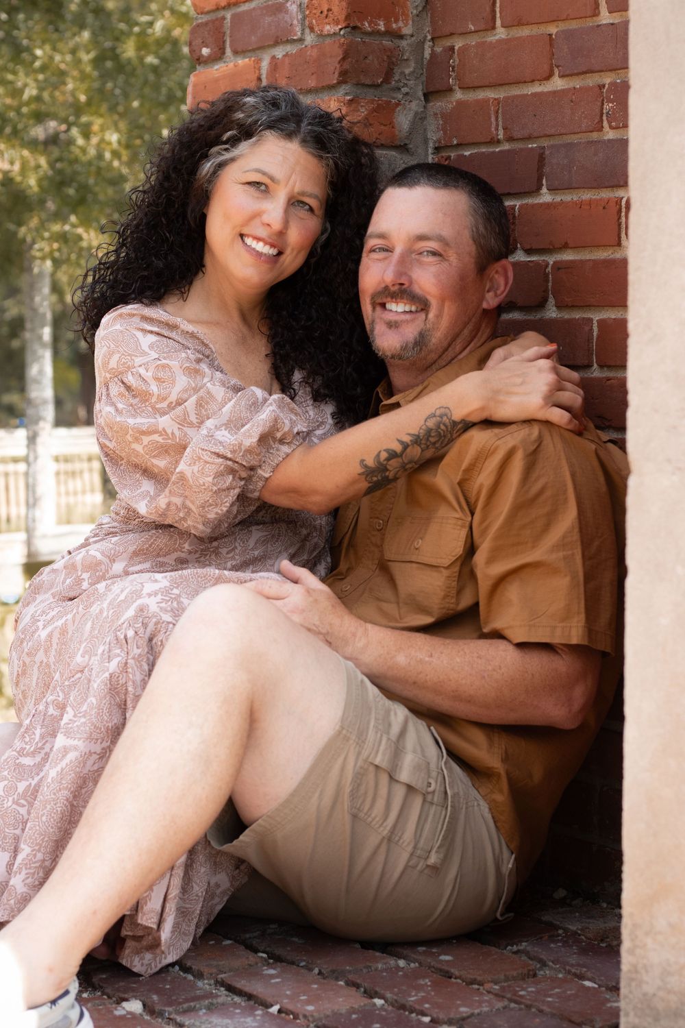 A couple in casual attire sits together against a brick wall, sharing a loving moment.