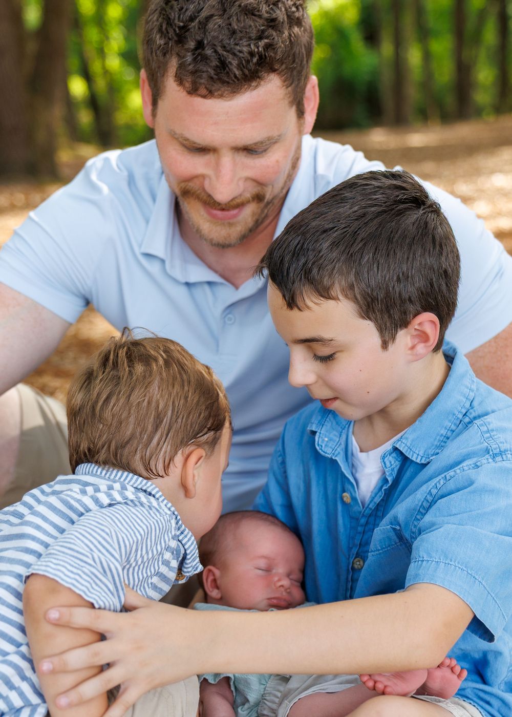 Father smiles as two young boys lovingly look at their newborn sibling outdoors in a sunlit forest setting.