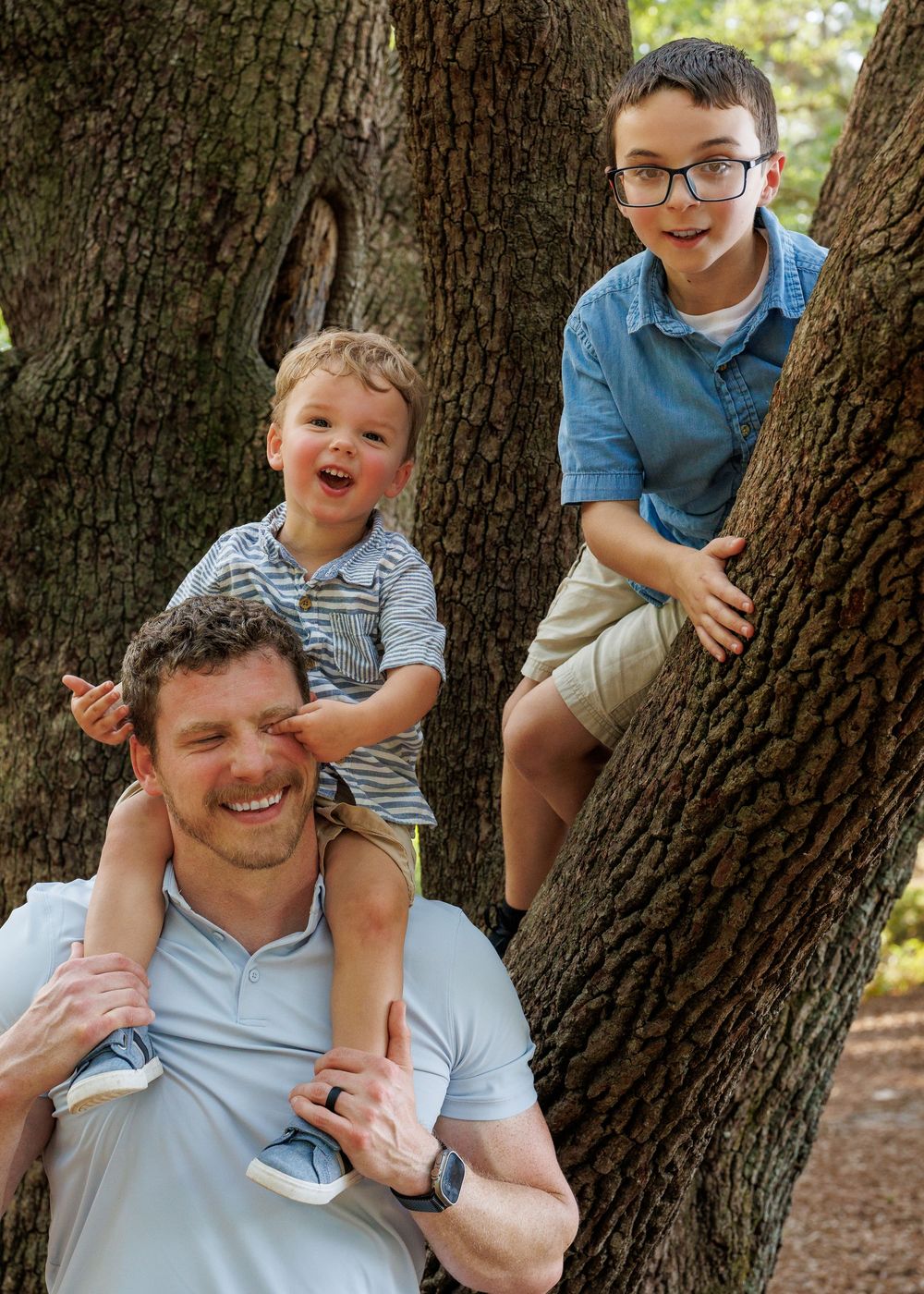 A smiling man holds a toddler on his shoulders while an older boy wearing glasses climbs a tree outdoors.