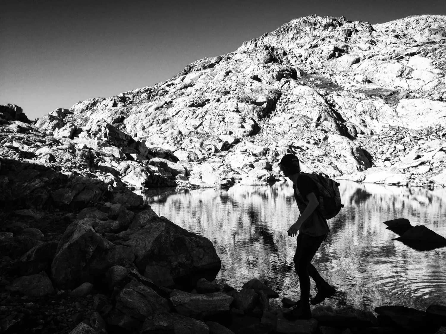 Silhouette d’un randonneur traversant des rochers près d’un lac alpin en noir et blanc, avec un reflet net dans l’eau calme.