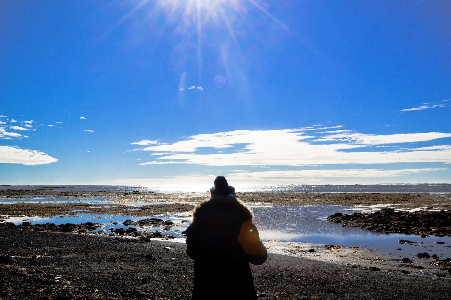 Silhouette d’une personne de dos observant l’océan sous un soleil intense, avec une lumière dure et un ciel bleu traversé de nuages.