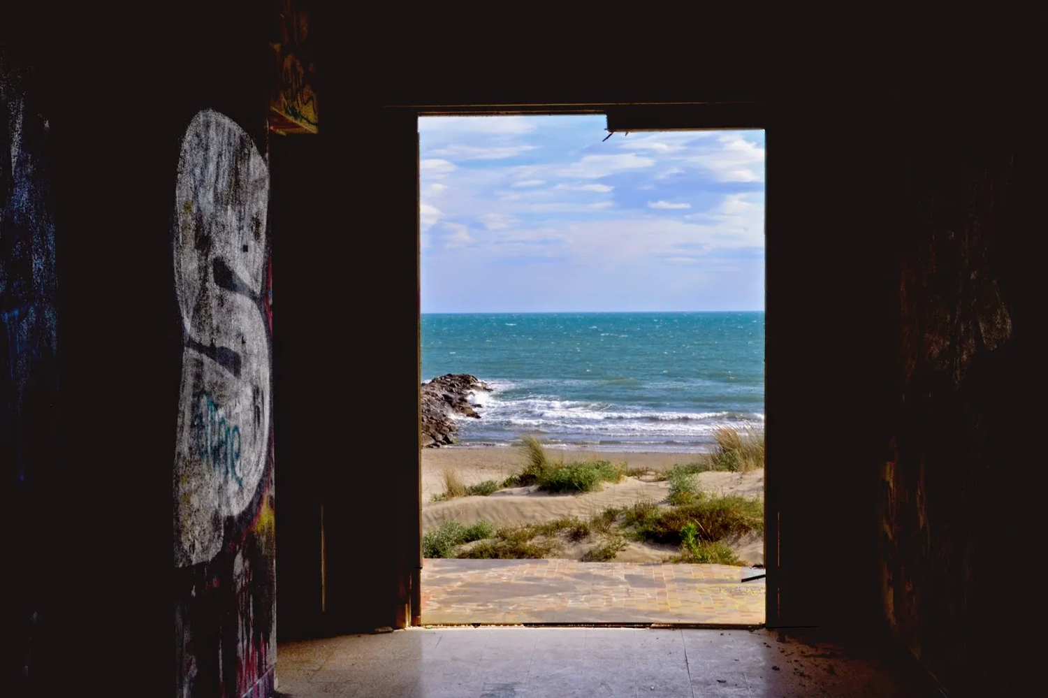 Cadrage sur une plage et l’océan vus depuis l’intérieur sombre d’un bâtiment abandonné, encadré par des murs tagués.