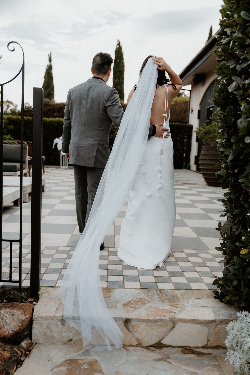 Elegant bride and groom standing on checkered stone steps with flowing white veil and cypress trees in the background.