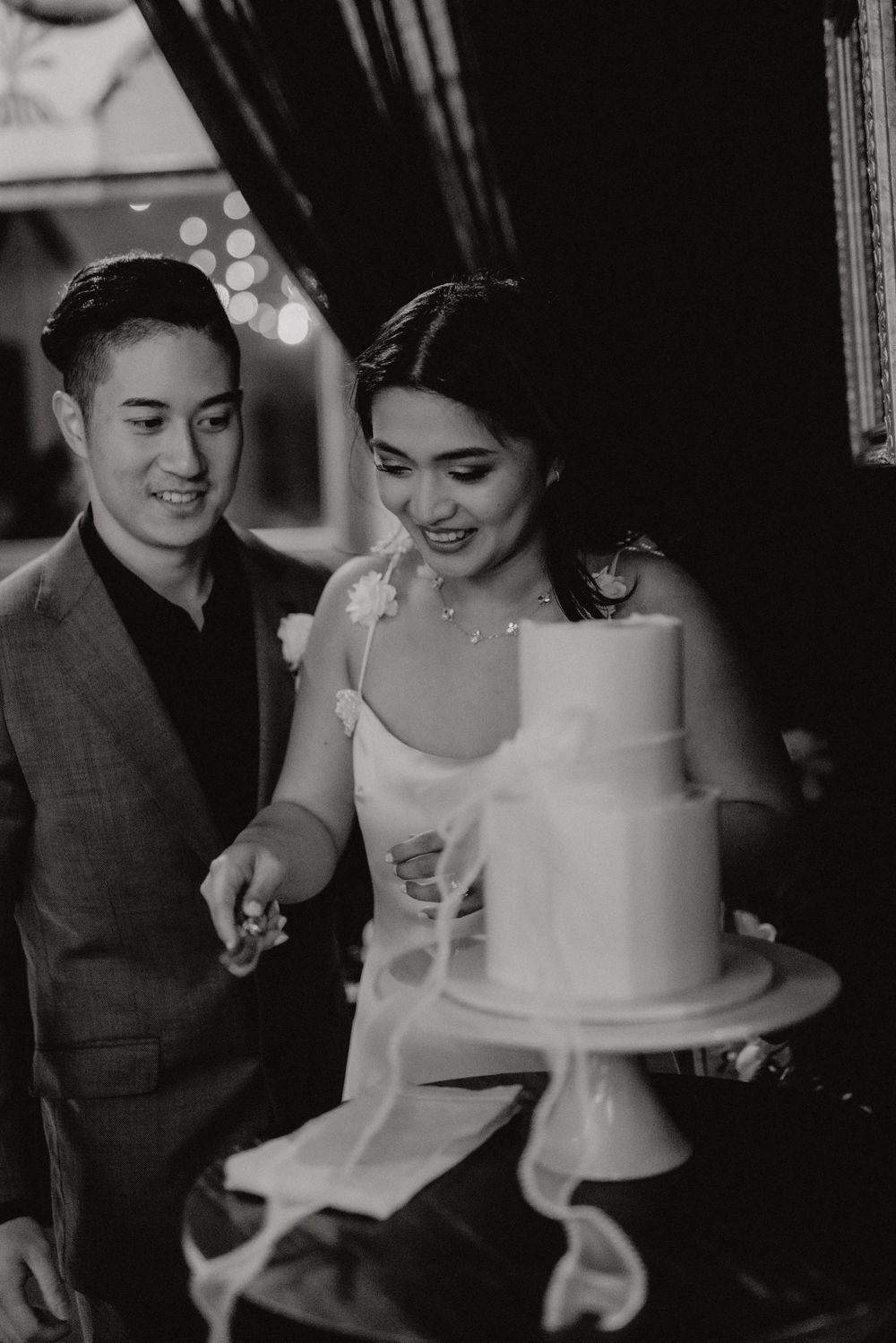 A couple in wedding attire smiling while cutting their elegant white tiered wedding cake together in dim romantic lighting.