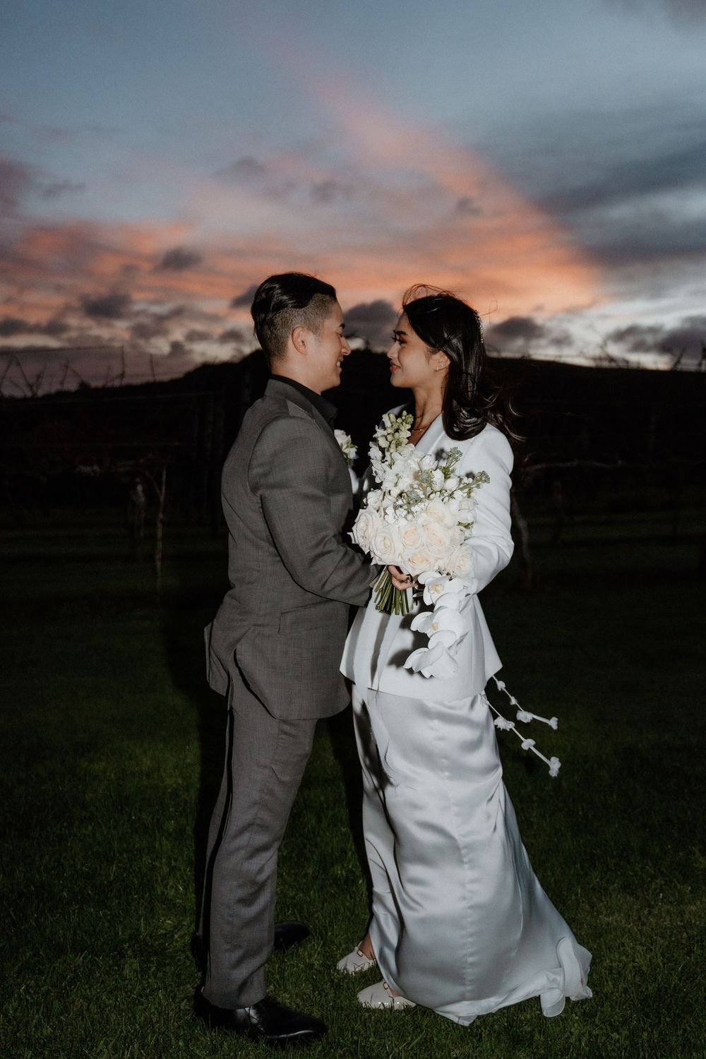 A couple in wedding attire share an intimate moment at sunset in a grassy field with dramatic cloudy sky.