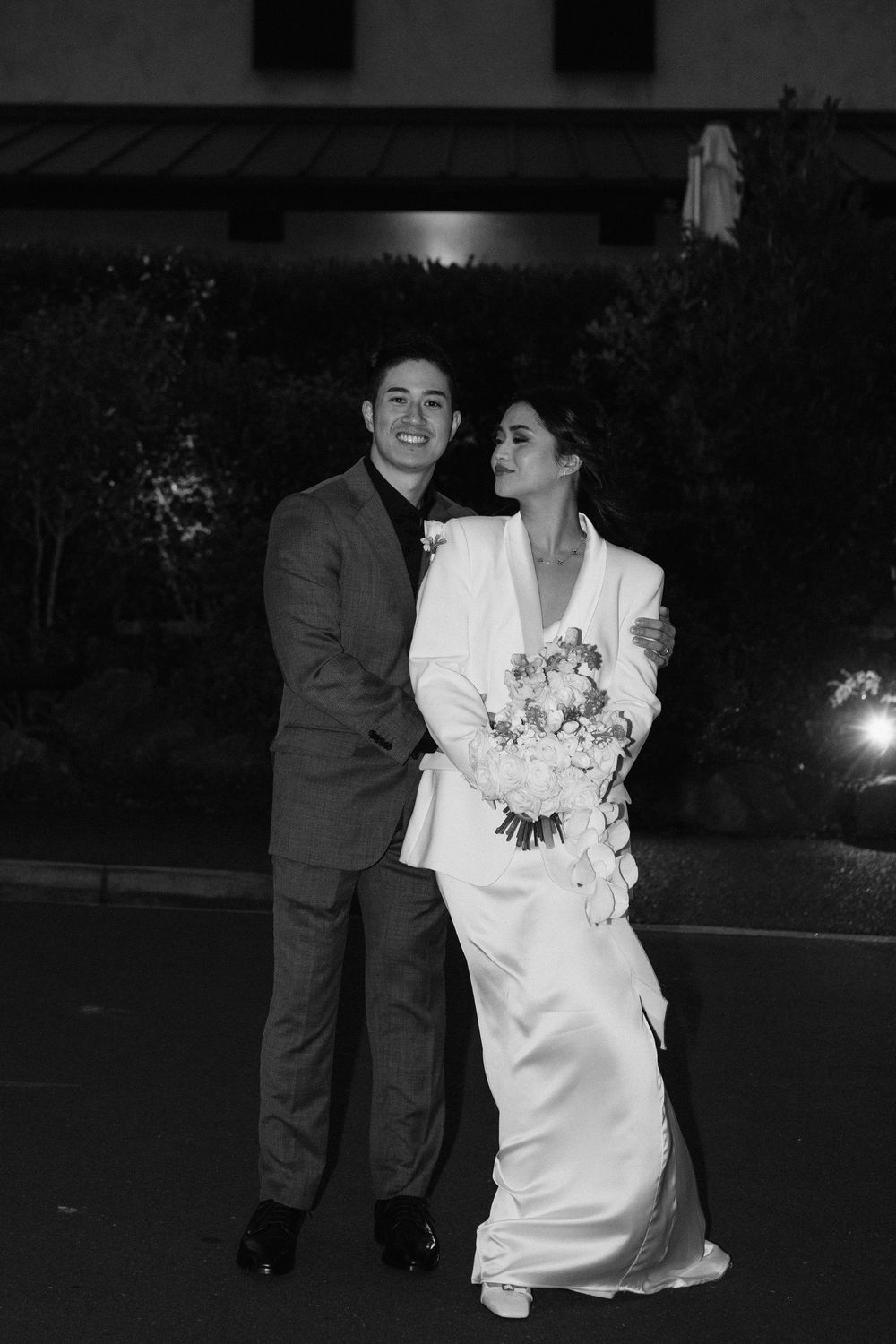 A black and white wedding photo showing a couple embracing outdoors at night, with the bride in a white gown holding flowers.