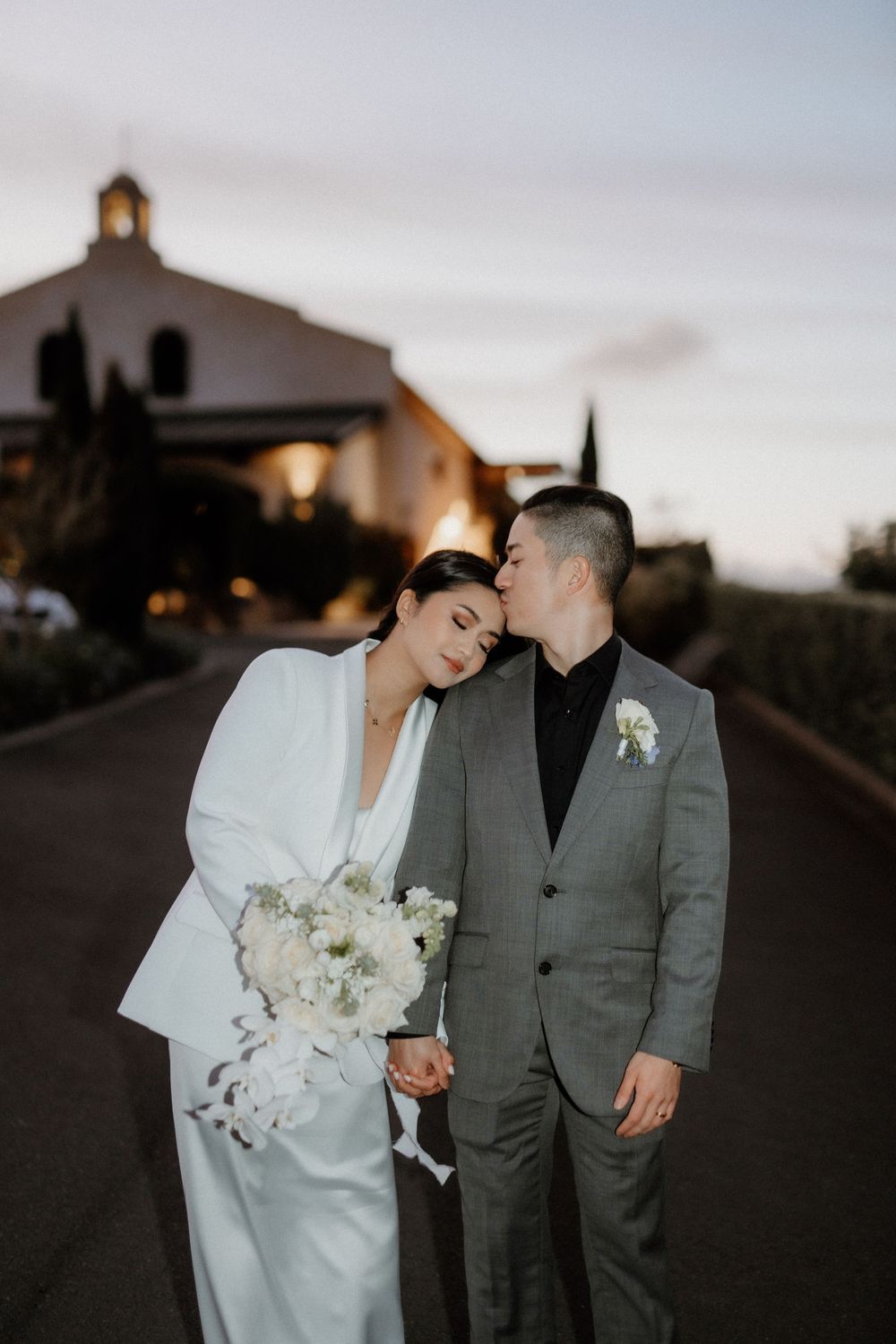 A wedding couple shares an intimate moment on a quiet road at dusk with a church building in the background.