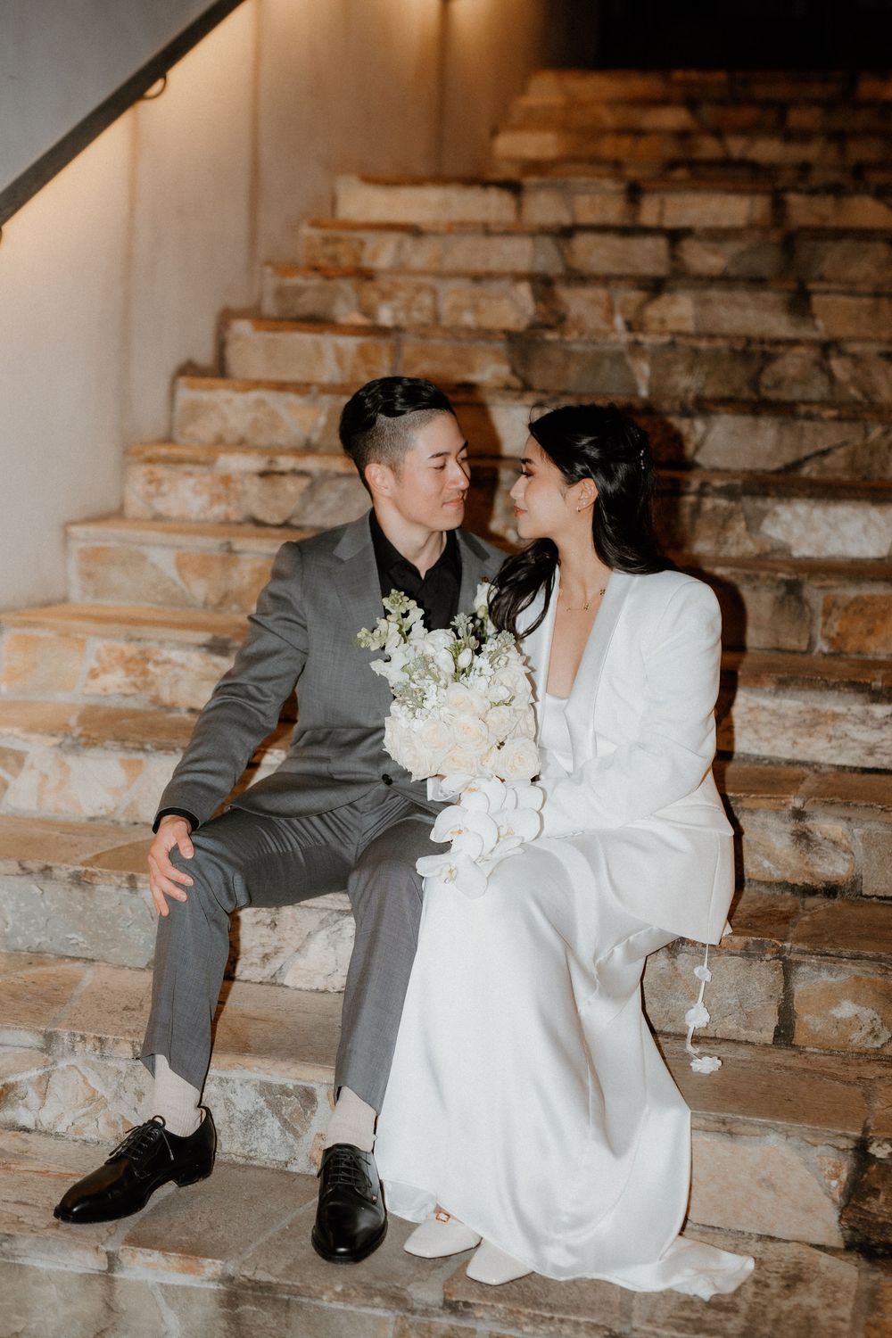 Elegant couple in wedding attire sitting on rustic stone stairs with white floral bouquet at romantic venue.