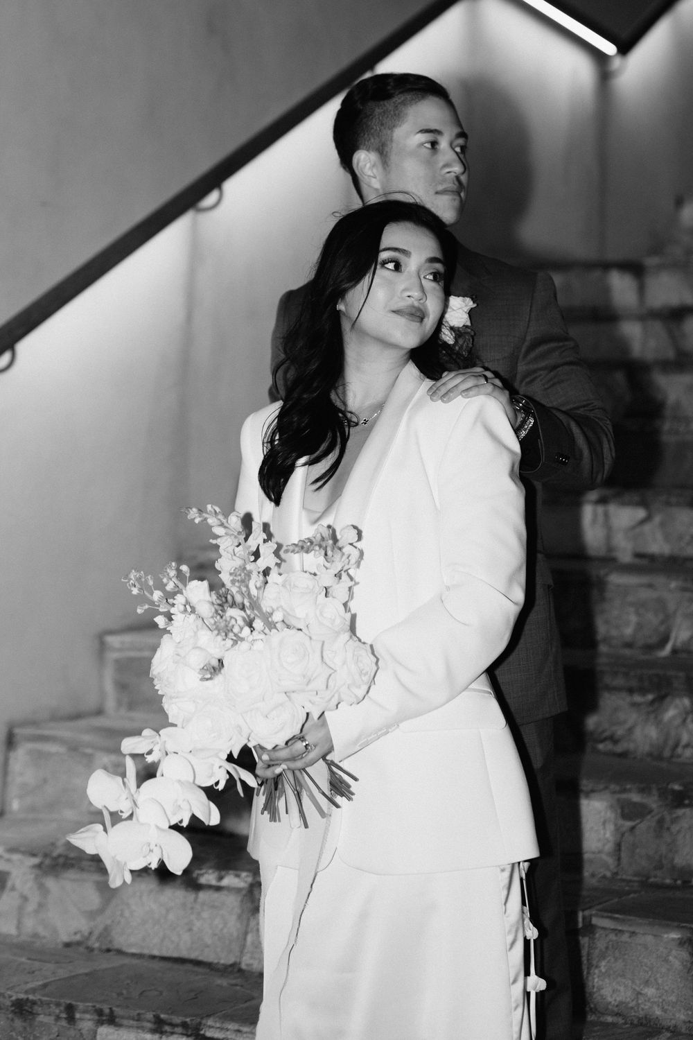Black and white photograph of a couple on a staircase, with the bride in a white dress holding a bouquet of flowers.