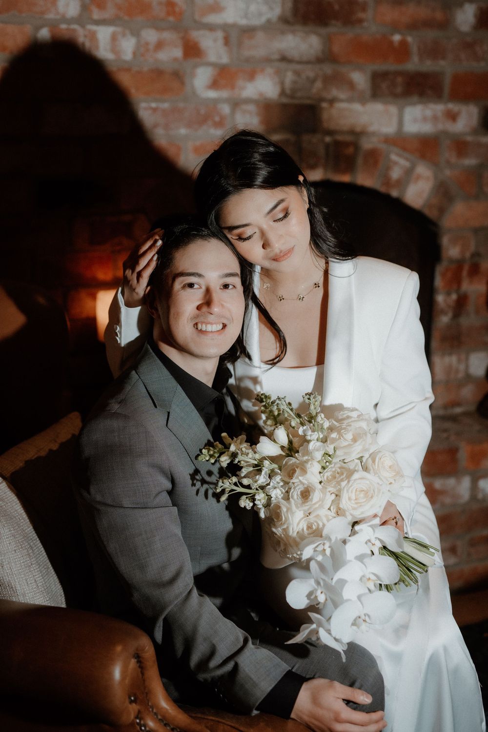Romantic wedding portrait set against rustic brick wall with dramatic lighting and white floral bouquet.