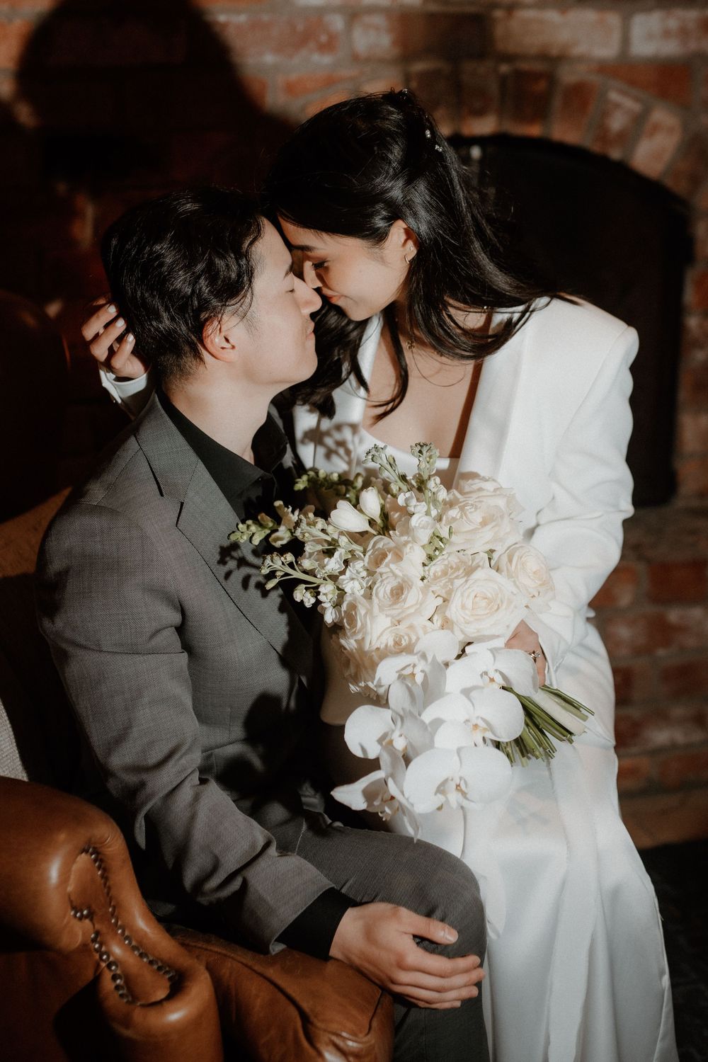 A romantic wedding moment captured near a brick wall with white orchids and roses in the bride's bouquet.