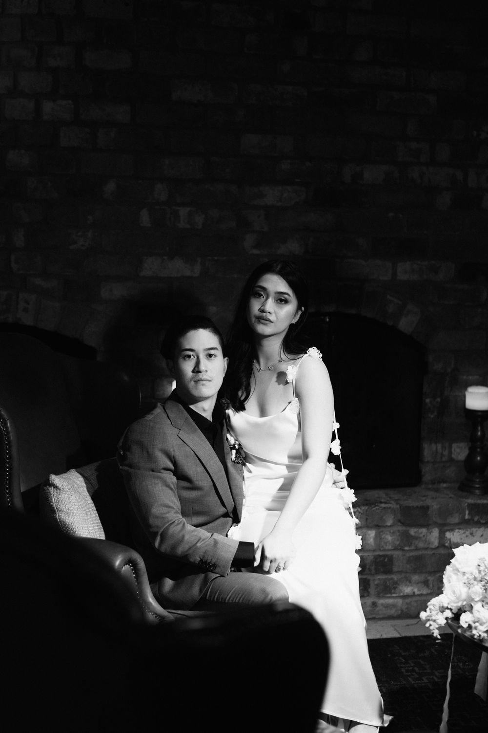 A romantic black and white wedding portrait shows a couple in formal attire posing elegantly against a brick wall backdrop.