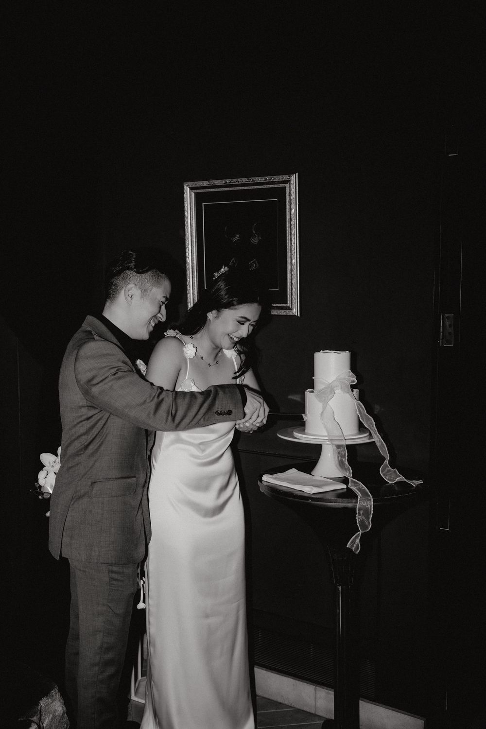 A couple in formal attire cuts their elegant white wedding cake together in a dimly lit room during their reception.