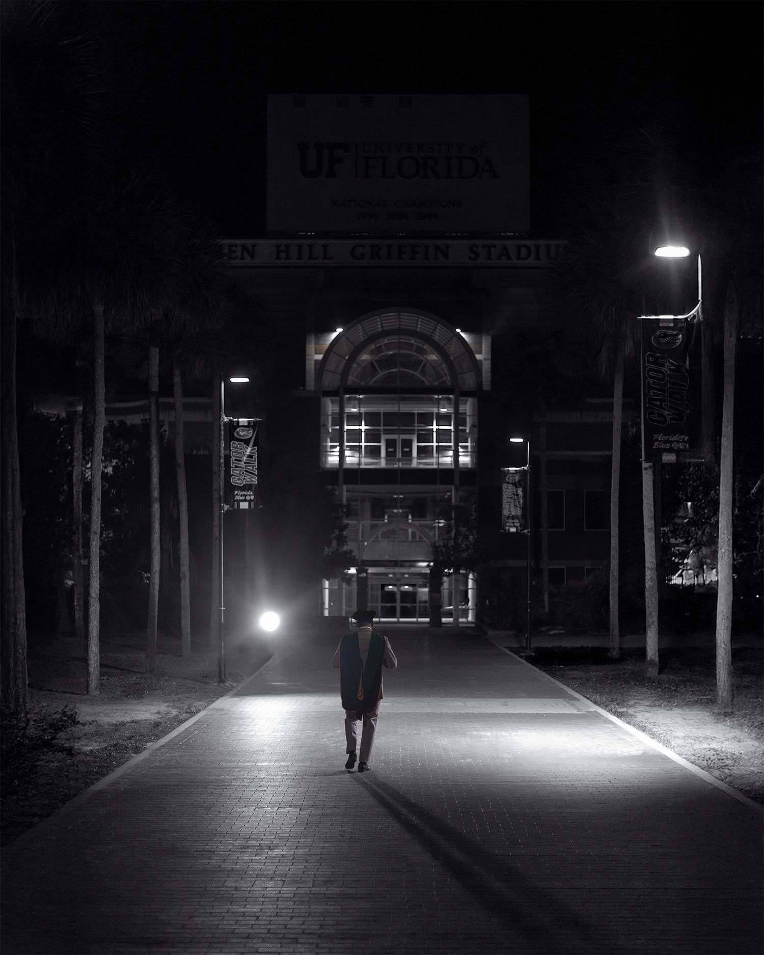 A solitary figure walks along an illuminated path towards an entrance of UF Plaza at night.