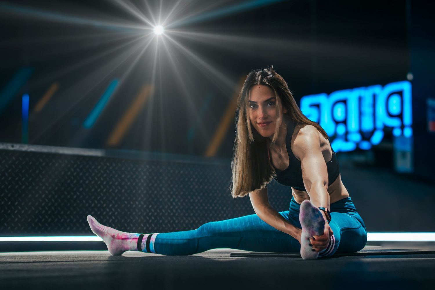 A woman in athletic wear performs a deep stretch on a dark floor under dramatic lighting.