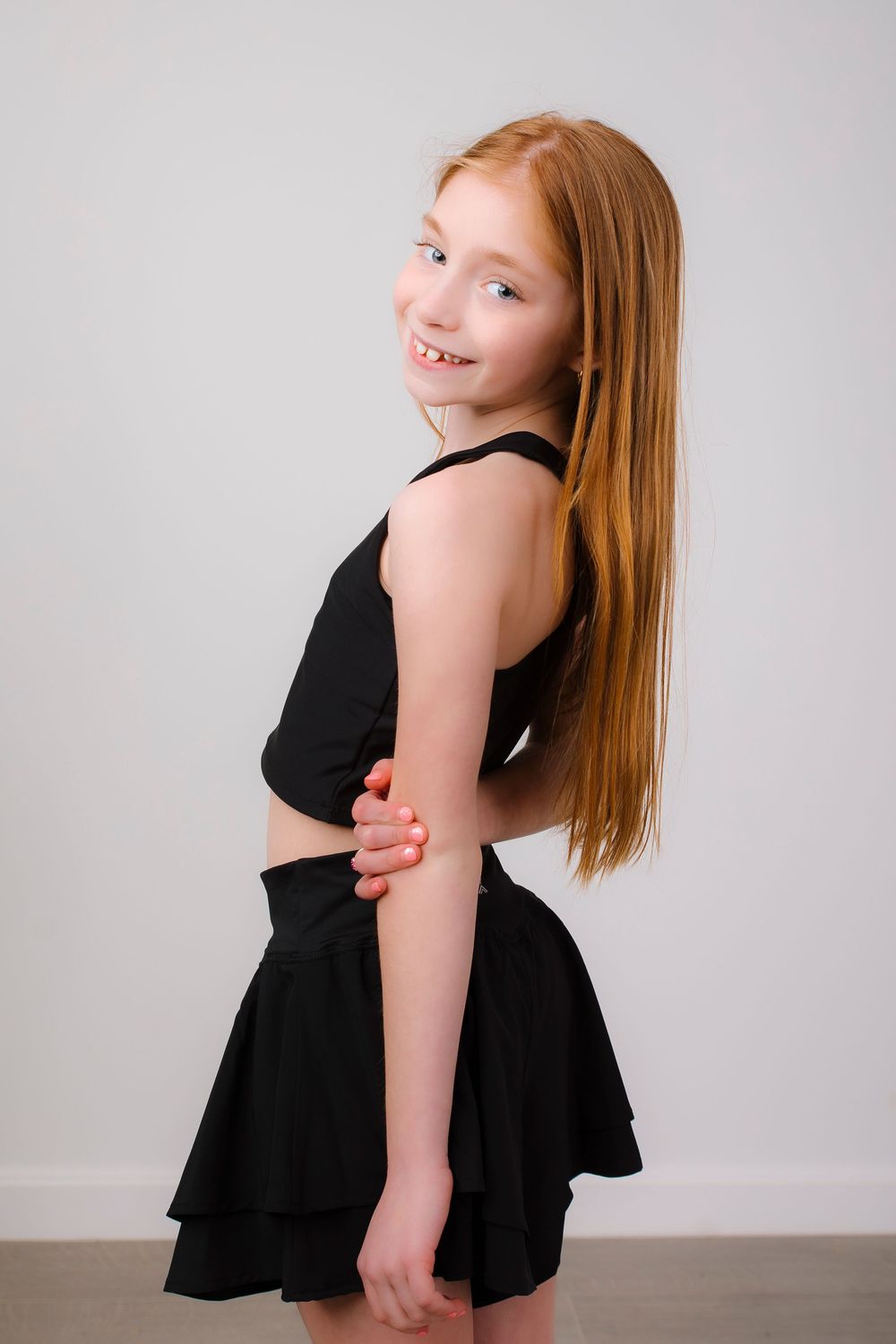 Young dancer in black two-piece outfit poses against gray background showing graceful movement.