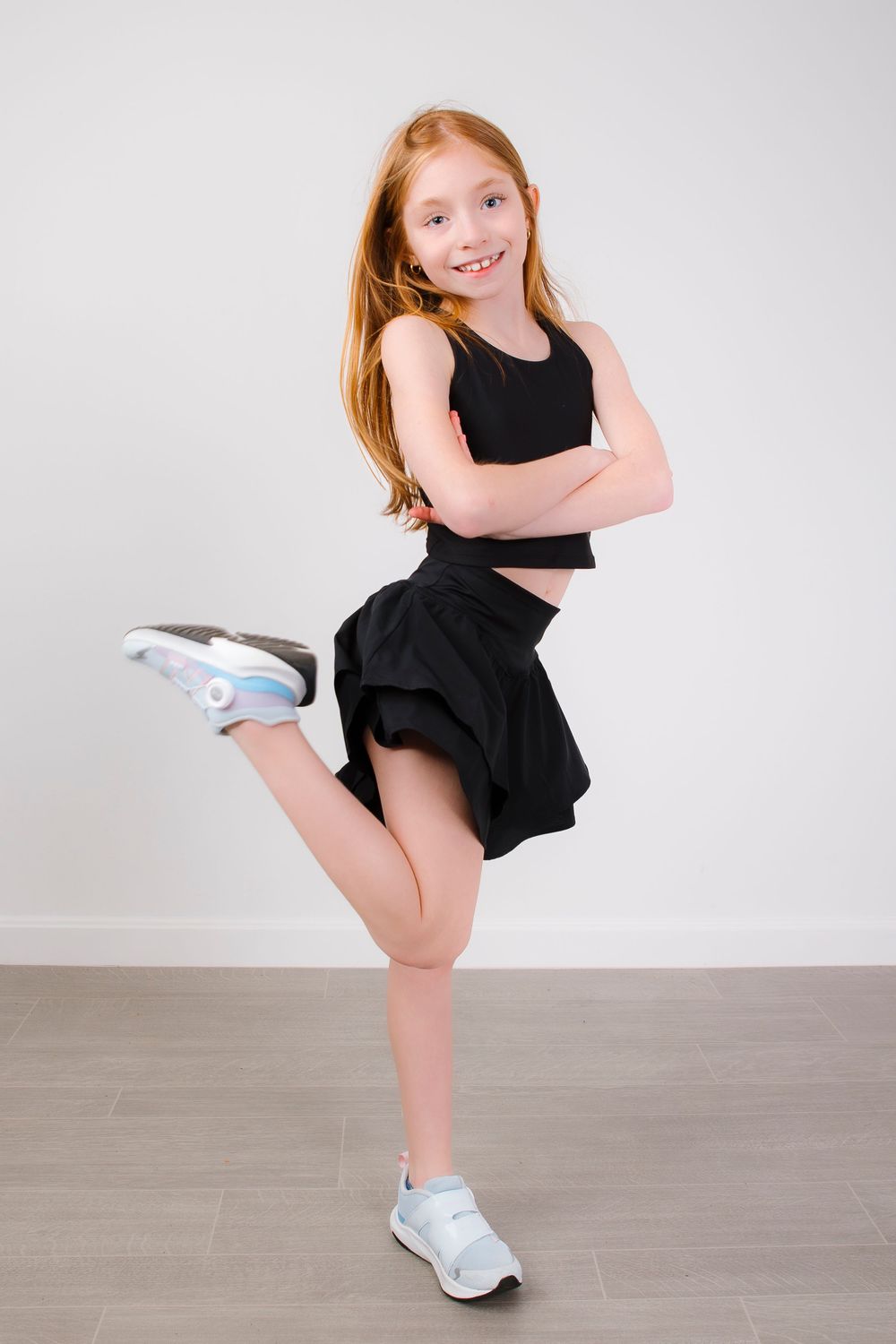 Young dancer in black outfit and white sneakers strikes energetic pose against light studio background.