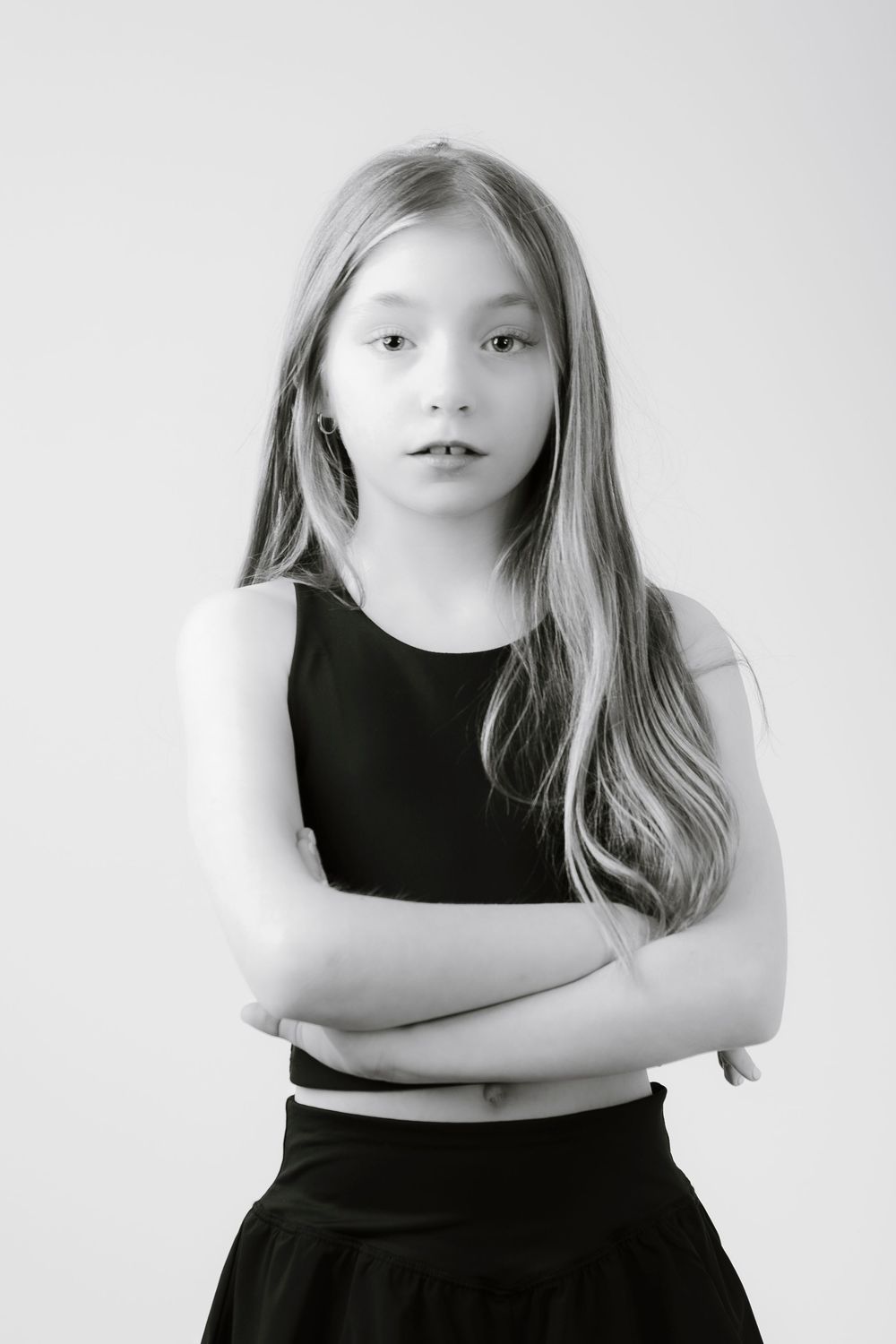 Black and white portrait of a young person with long hair wearing a black outfit standing with arms crossed.