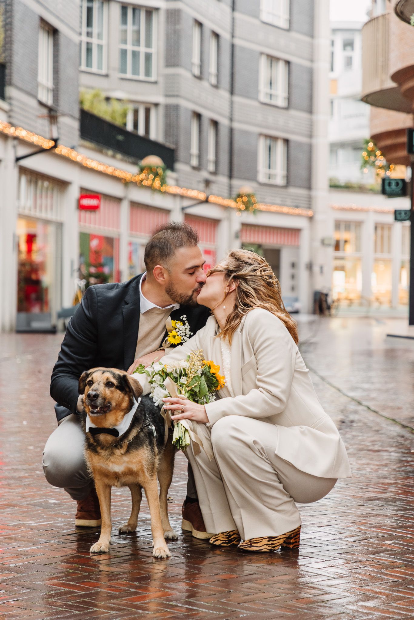 A couple shares a romantic kiss on a rainy cobblestone street with their adorable dog sitting beside them.
