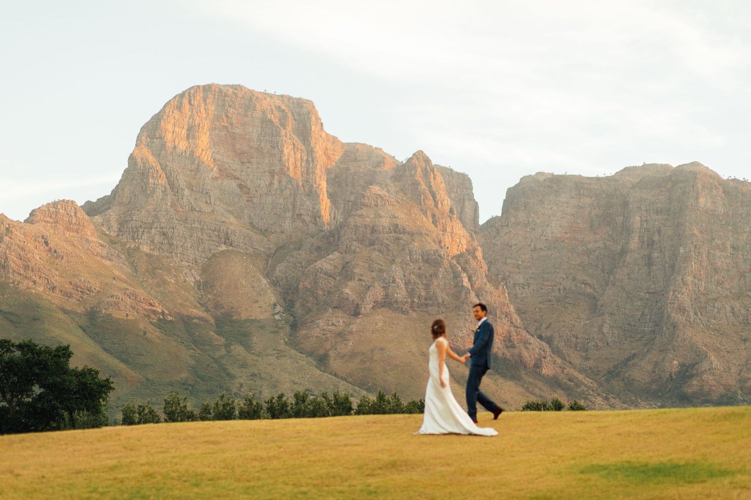 Two figures in formal attire walk across a green lawn with majestic mountain peaks glowing in the sunset behind them.