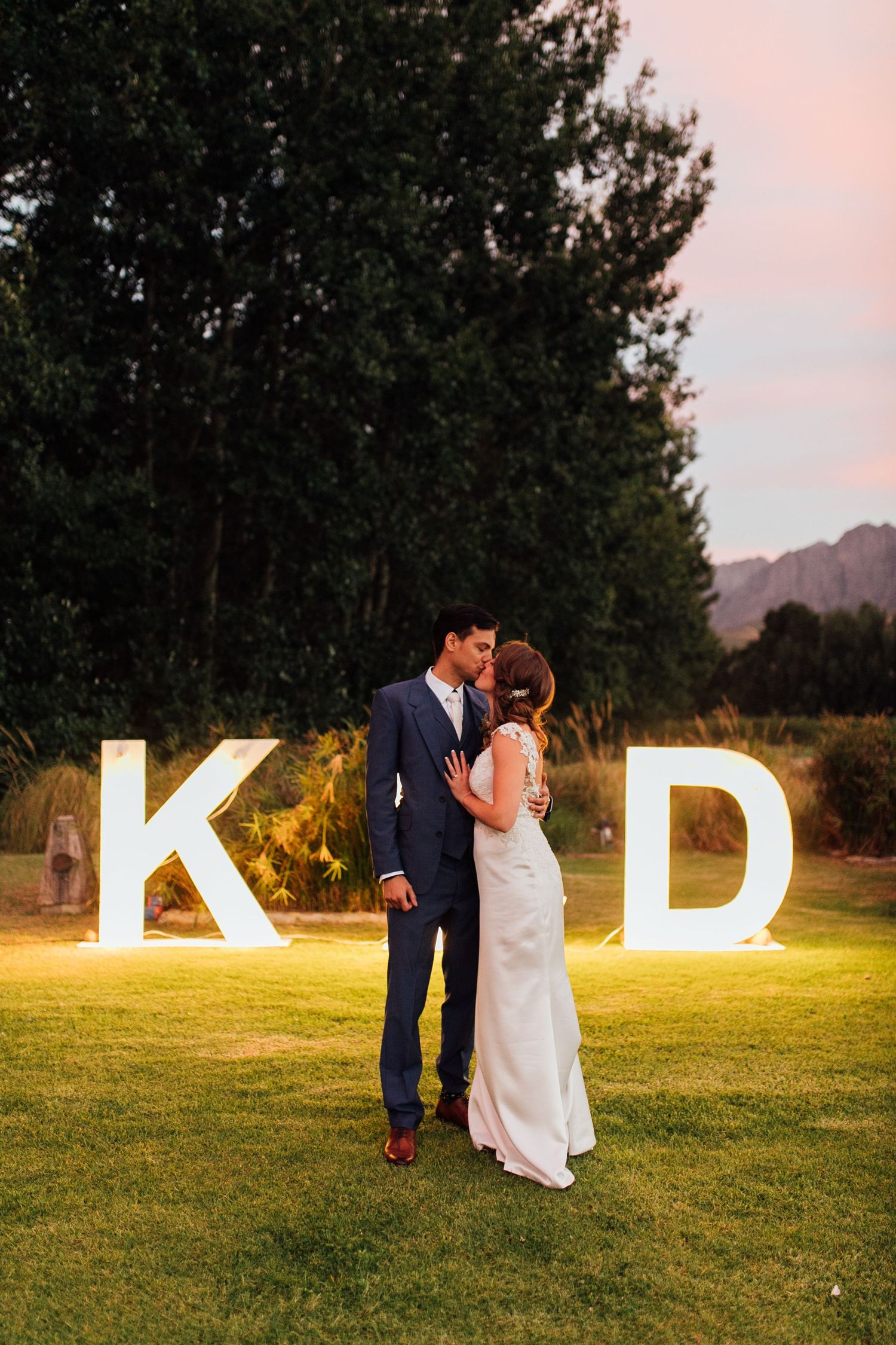 Couple embraces on a grassy lawn at sunset with illuminated K & D letters displayed behind them in a mountain setting.