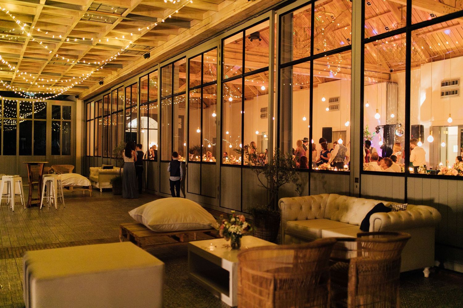 Modern restaurant interior with glass walls, warm lighting, and cozy lounge seating on a patio area at night.
