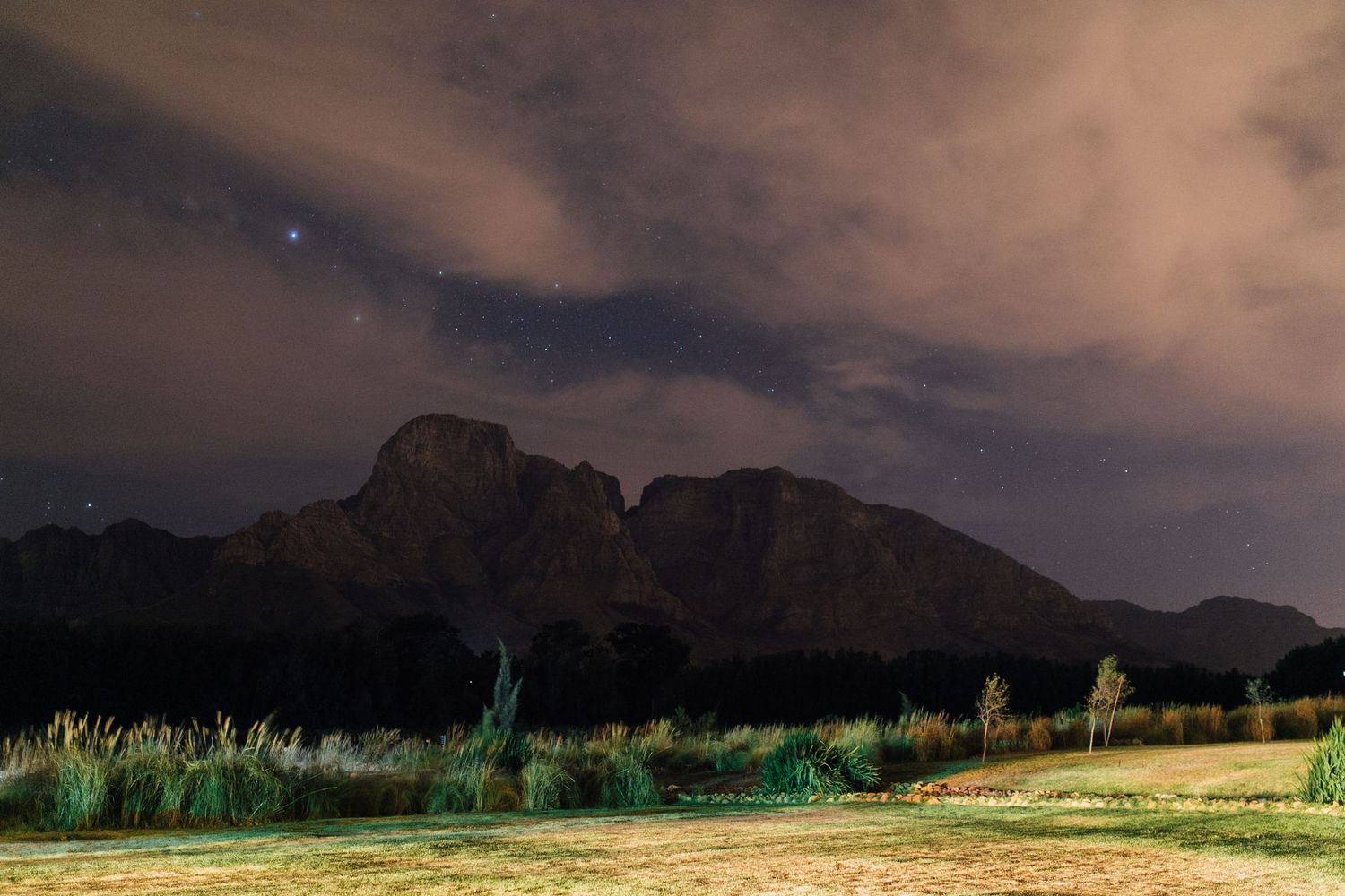A nighttime landscape of rugged mountains under cloudy skies with illuminated green vegetation in the foreground.