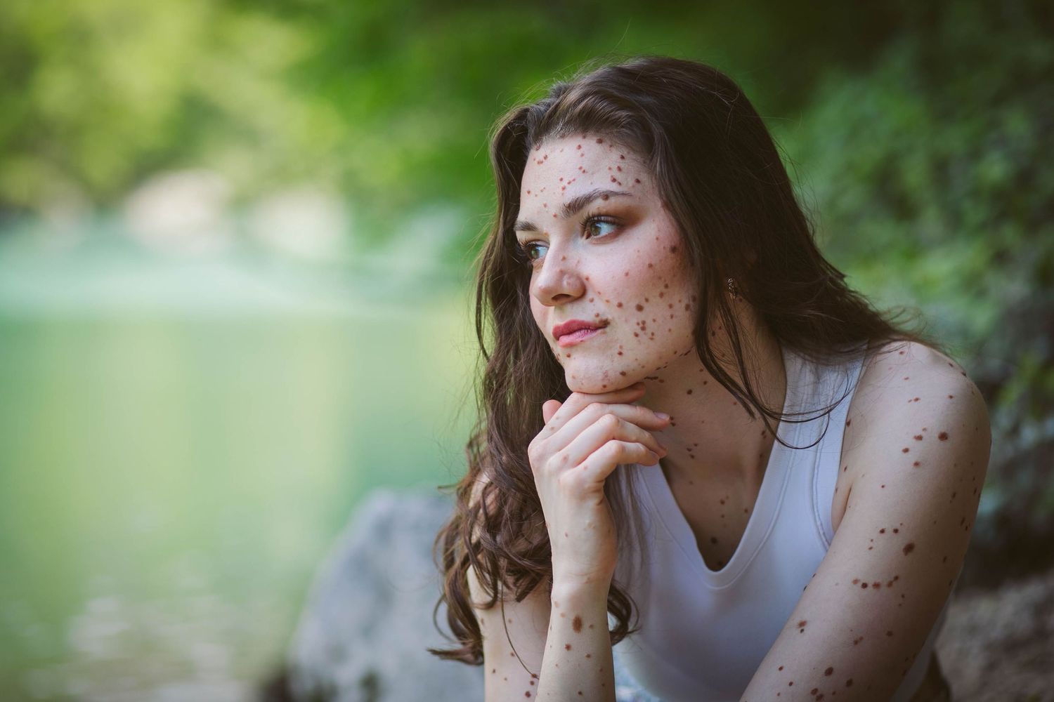 Thoughtful portrait in white tank top sits by lake with soft bokeh nature backdrop and freckled skin.